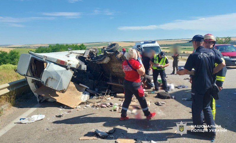 A damaged white van lies overturned on its side along a rural roadside as emergency responders, including police officers and a medic in a red vest, gather at the scene amid scattered debris.
