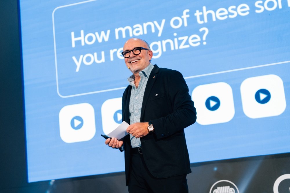 A smiling, bald man with glasses in a dark blazer speaks on stage, holding a paper, before a screen reading "How many of these... you recognize?" with play-button icons.