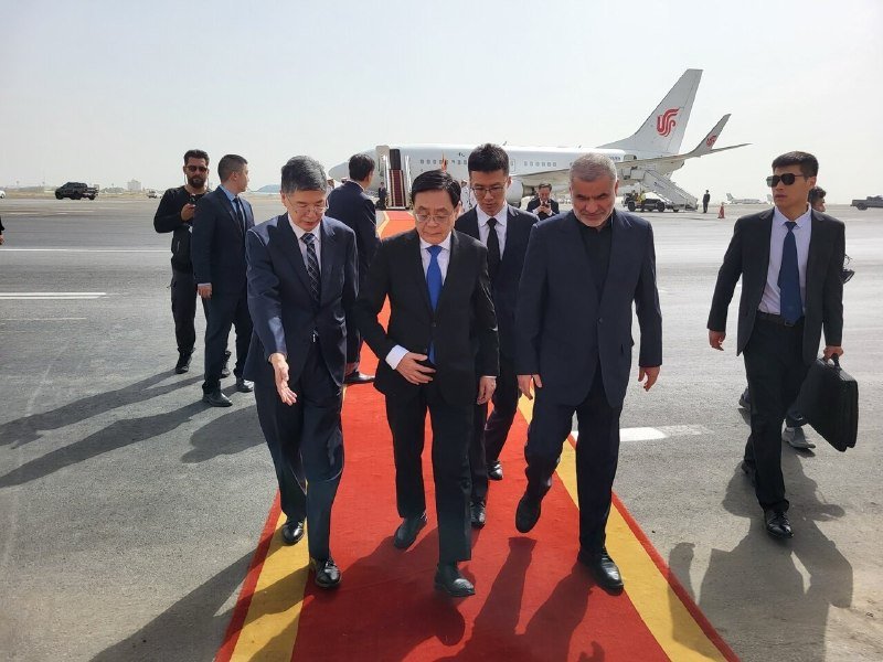 A group of suited men walk along a red and yellow carpet on an airport tarmac, with a white passenger airplane parked behind them.