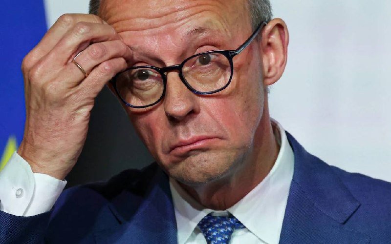 A man in a blue suit, blue tie, and glasses rests his hand against his forehead with a serious expression.