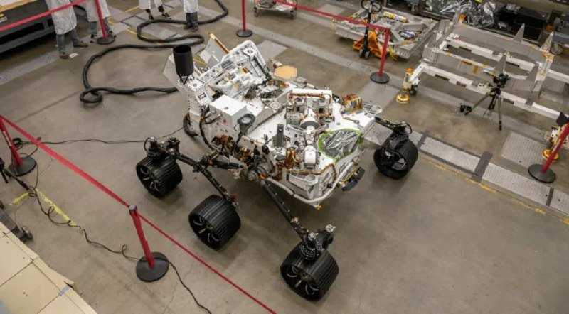 A six-wheeled robotic rover with exposed mechanical and electronic components sits on a concrete floor behind red stanchions in a facility, with technicians visible in the background.