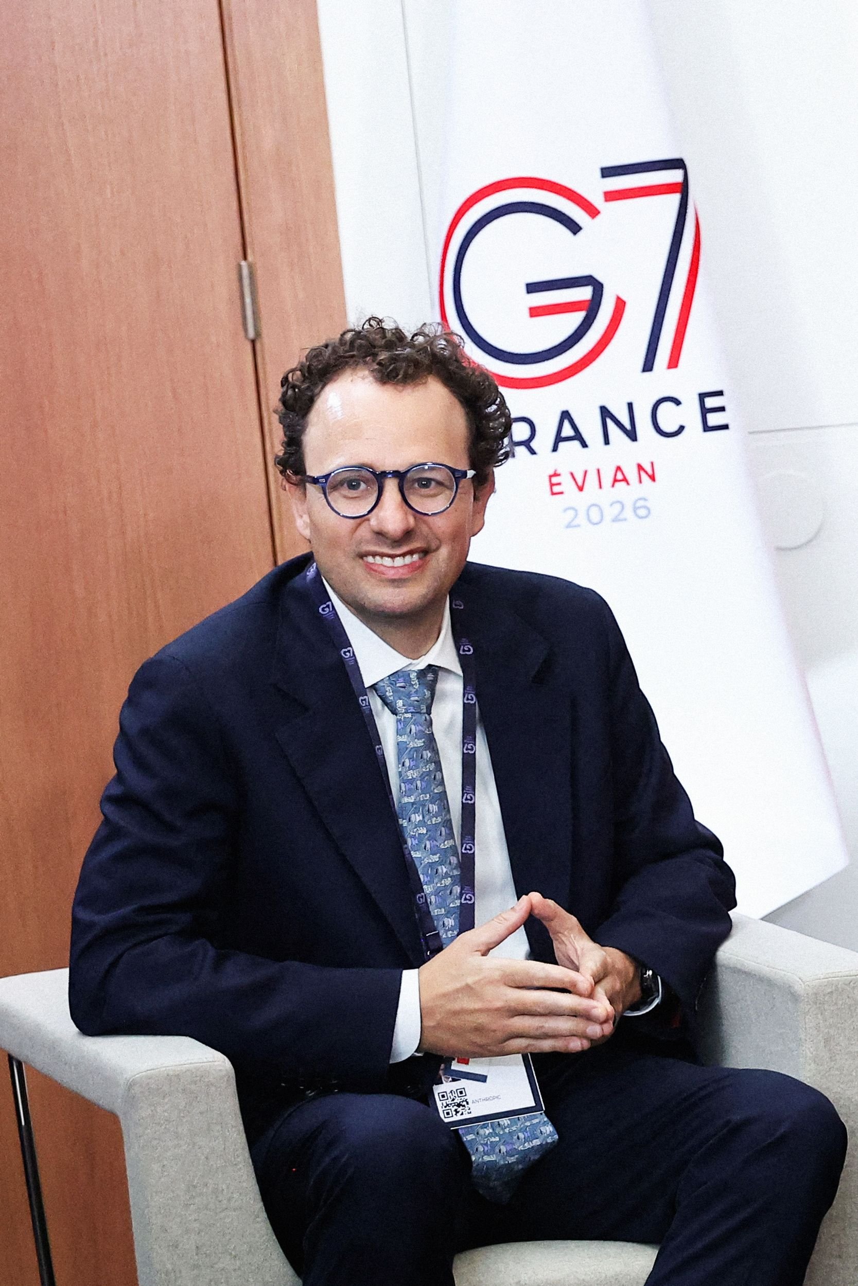 A smiling man in a dark suit, glasses, and lanyard sits in a chair with hands clasped, in front of a "G7 FRANCE ÉVIAN 2026" backdrop.