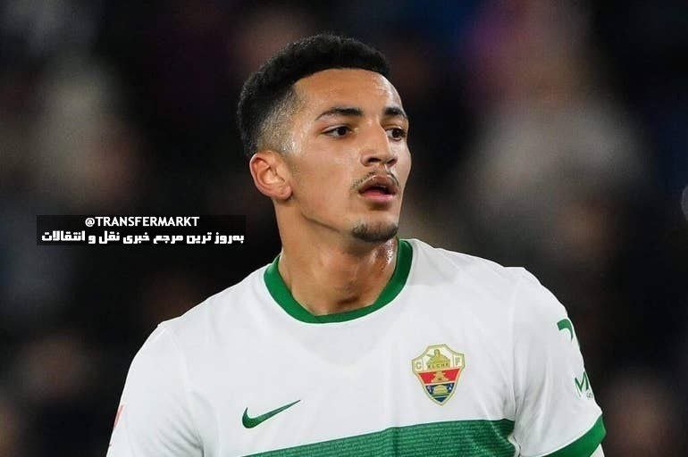 A soccer player with short dark hair and a goatee looks to the side while wearing a white jersey with green trim and a team crest.