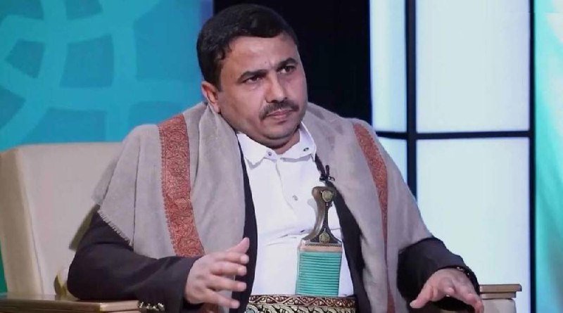 A man wearing a white shirt and a beige shawl with red trim sits on a chair during what appears to be a televised interview, gesturing with his hands.