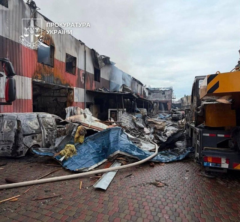 Burned-out building debris, charred vans, and a yellow crane truck sit on a brick-paved lot, with an emblem and Cyrillic text "ПРОКУРАТУРА УКРАЇНИ" overlaid in the top-left corner.