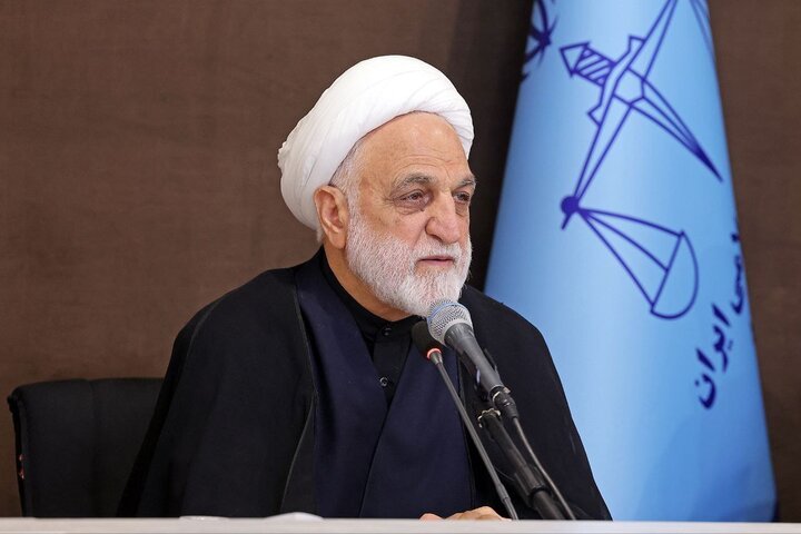 A white-bearded man wearing a white turban and black clerical attire speaks into a microphone, positioned before a light blue flag featuring Persian script and an emblem.