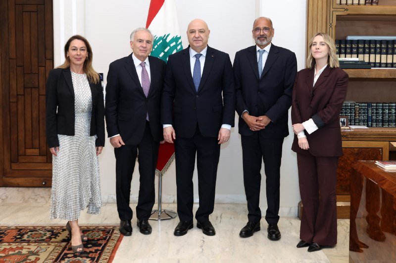 Five formally dressed individuals stand in a row in an ornate wood-paneled room, with a Lebanese flag displayed behind them.