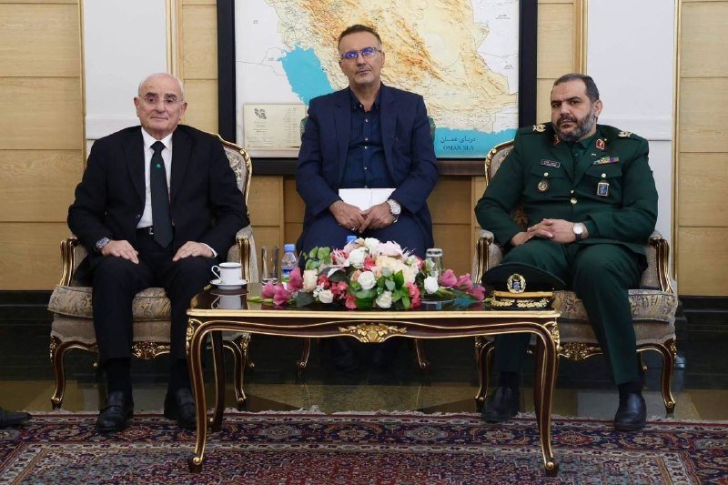Three men sit posed in ornate chairs behind a floral-topped coffee table, with a regional map displayed on the wall behind them.