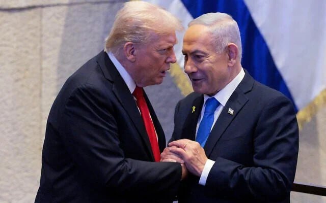 Two men in dark suits shake hands and lean close together, one wearing a red tie and the other a blue tie, with a blue-and-white flag visible in the background.