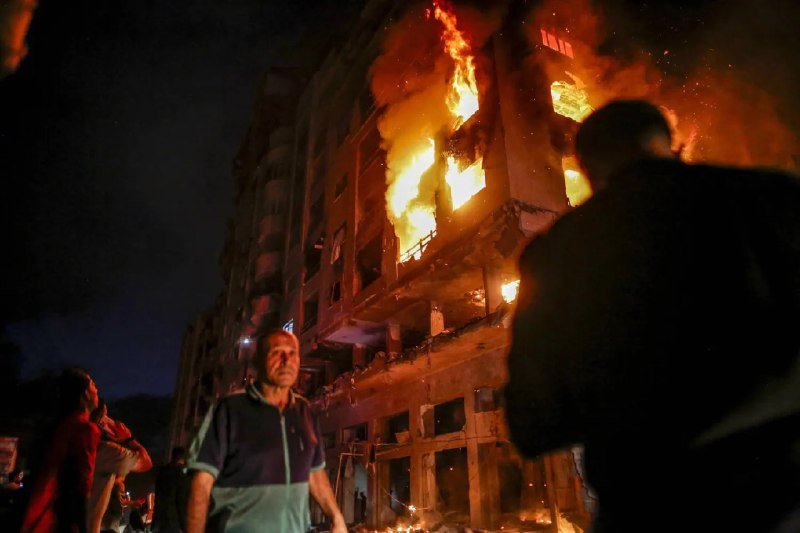 A multi-story building engulfed in flames and thick smoke at night, with several people gathered in the foreground watching the fire.