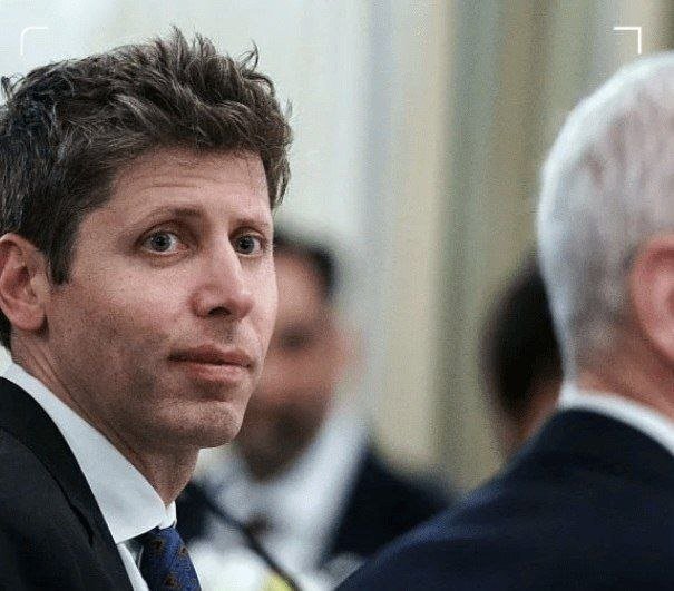 A man in a dark suit and blue tie looks over his shoulder toward an older man with white hair, set against a blurred indoor background.