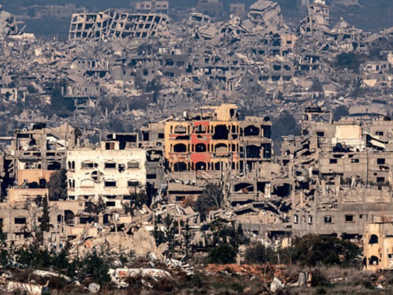 Extensive rubble and heavily damaged multi-story buildings stretch across a wide urban landscape under a hazy sky.