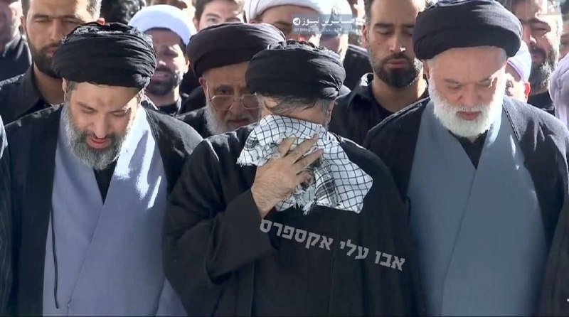 A man wearing a black turban and keffiyeh is embraced by others in black clerical attire, with Hebrew text overlaid and "Tasnim News" watermark visible.