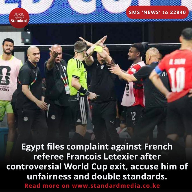 A referee in a yellow shirt holds up a yellow card as players in black and red kits argue intensely on a football pitch.