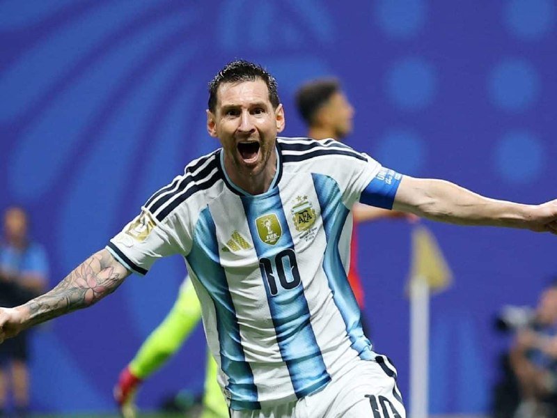 A soccer player wearing the number 10 Argentina jersey with black stripes celebrates with arms outstretched and mouth open during a match.