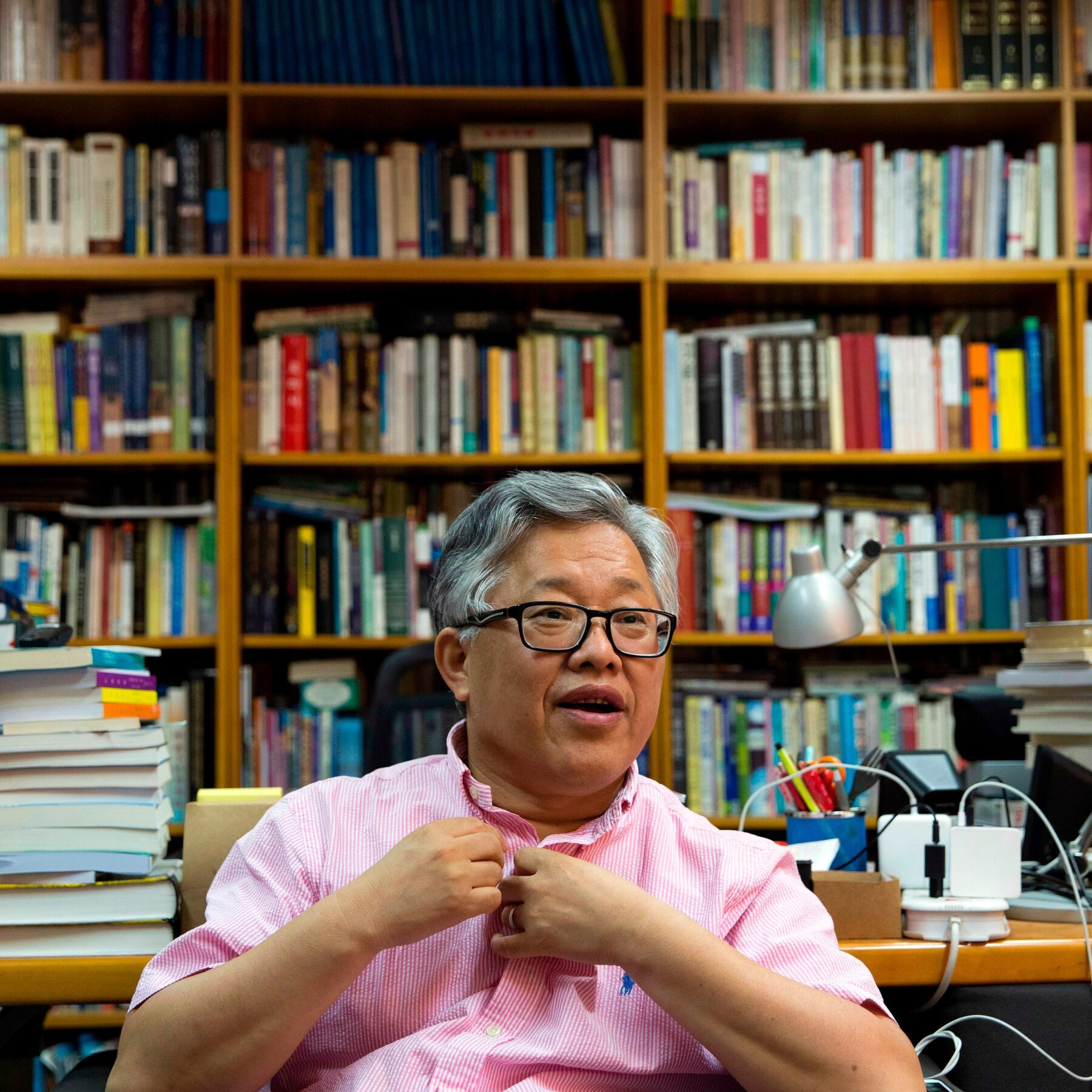 An older man with gray hair and glasses, wearing a pink striped shirt, sits in front of floor-to-ceiling bookshelves filled with books.