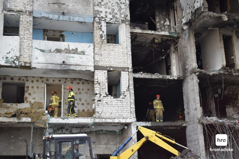 Firefighters in helmets and protective gear stand inside the gutted, shrapnel-pocked shell of a multi-story building, inspecting damage near a yellow excavator.