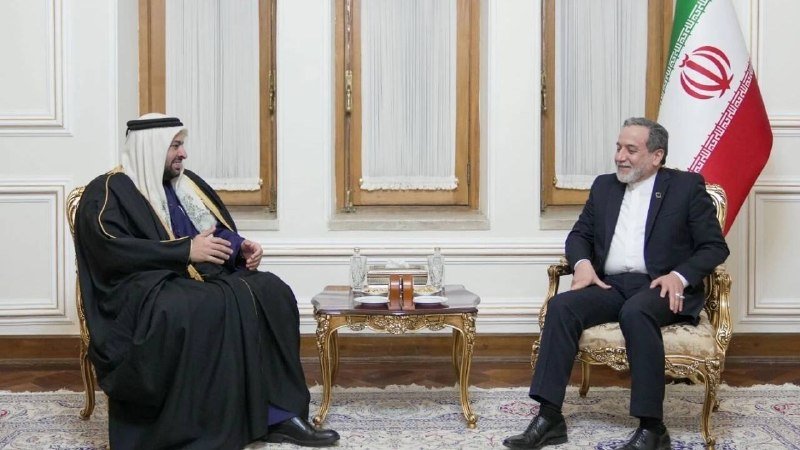 Two men in formal attire — one in traditional Arab dress, the other in a dark suit — sit in ornate chairs across a small table, with an Iranian flag visible behind them.