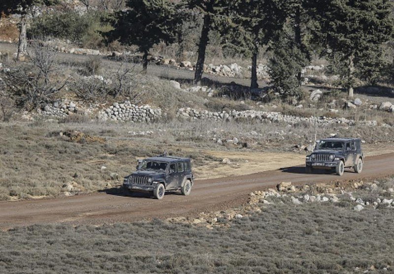 Two dark off-road vehicles drive along a dirt road through a rocky, tree-lined landscape with dry grass and stone walls.