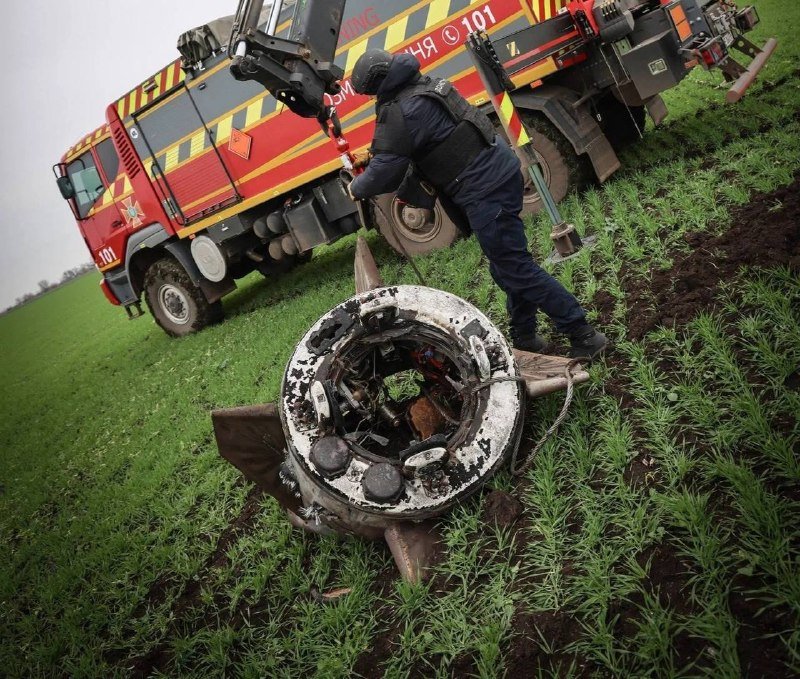 A bomb squad technician in tactical gear prepares a charred, spiked fragment of a destroyed munition next to a red fire/emergency truck marked "101" in a grassy field.