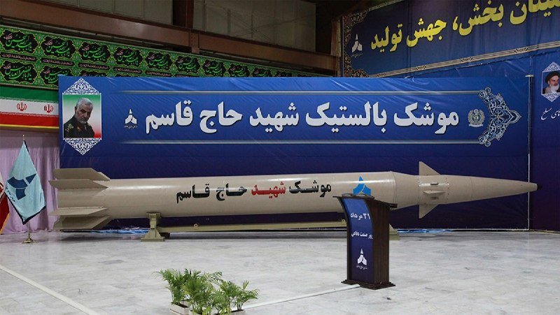 A large missile labeled "حاج قاسم" is displayed indoors beneath a blue banner with Persian text and a portrait of a man in military uniform.