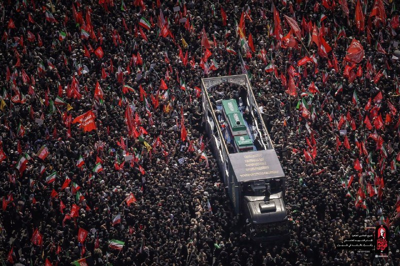 An elevated view shows a vehicle carrying a coffin surrounded by a massive crowd holding red and Iranian flags, with Persian text visible on banners.