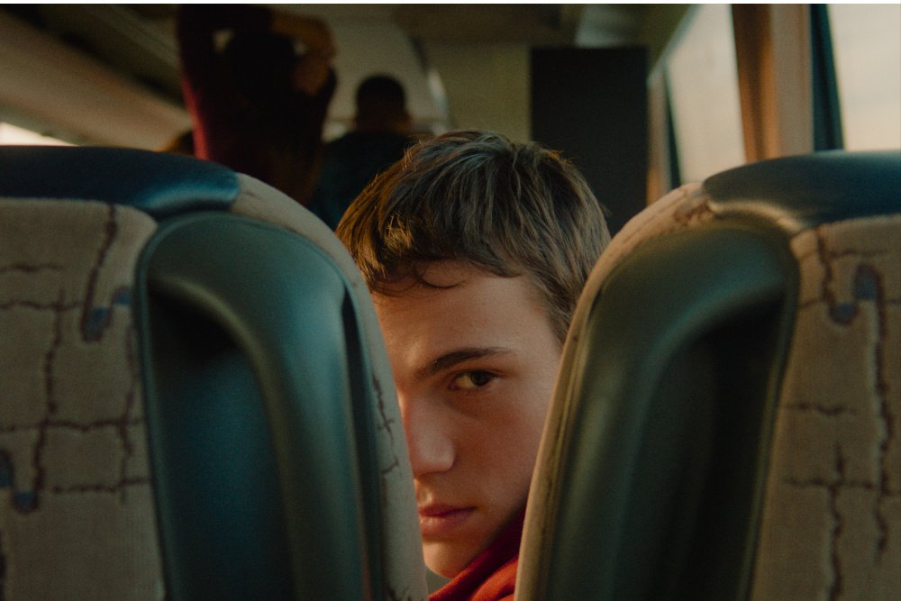 A young person with short brown hair peeks between two patterned bus seats, with other passengers visible in the blurred background.