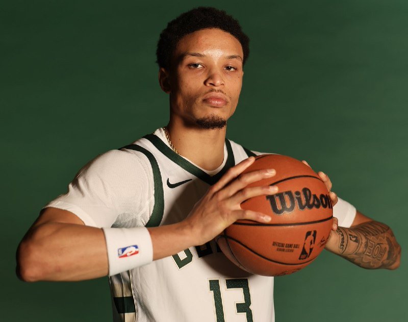 A young man in a white and green basketball jersey holds a Wilson basketball against a solid green backdrop during what appears to be a media photo session.