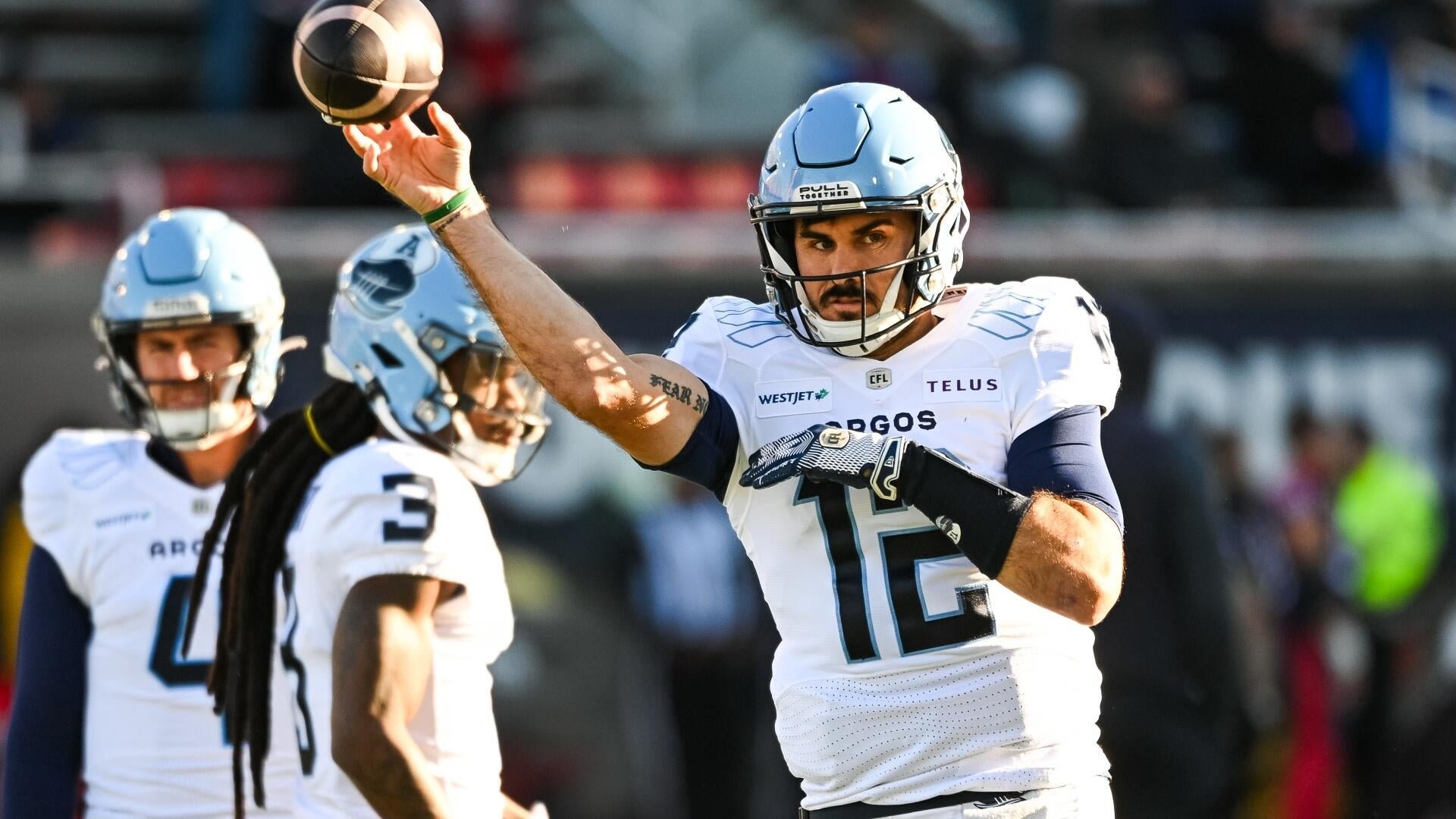 A football player wearing a white Argonauts jersey (#12) reaches out to catch a football with one hand during a game, with two teammates visible behind him.