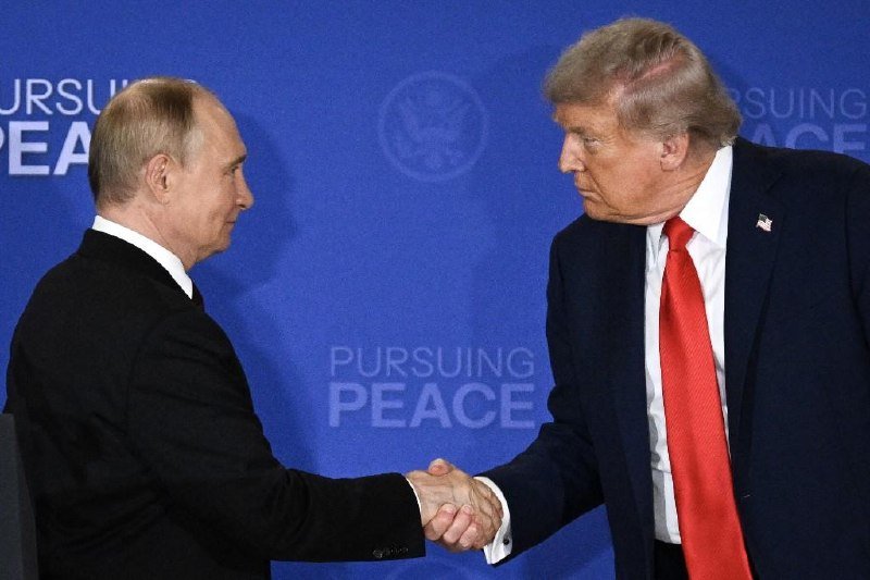Two men in dark suits shake hands in front of a blue backdrop displaying the words "PURSUING PEACE."
