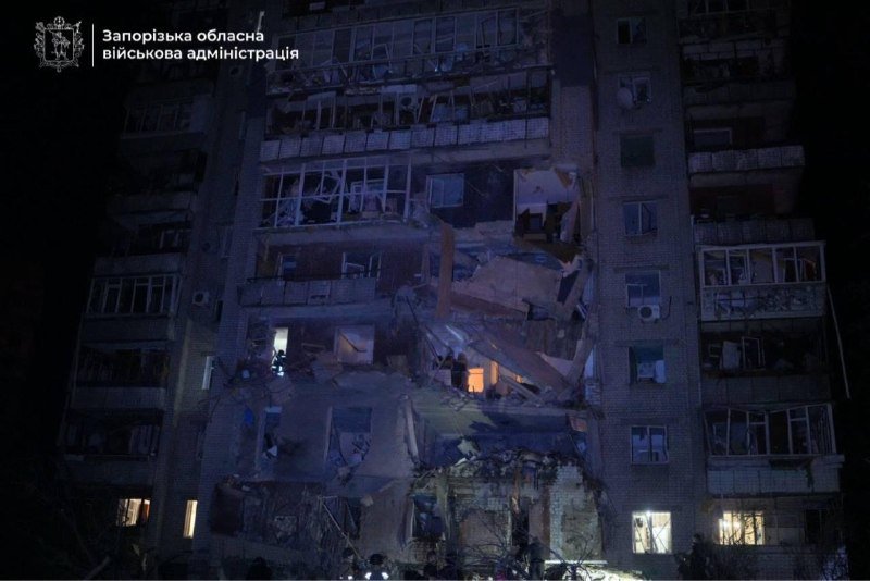 A nighttime image shows a multi-story residential building with extensive structural damage, shattered windows, and debris scattered across multiple floors, featuring a Ukrainian regional military administration logo in the upper left corner.