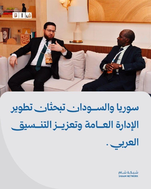 Two men in suits sit on a white sofa in a brightly lit room, conversing, with Arabic text and the "Shaam Network" logo displayed below.