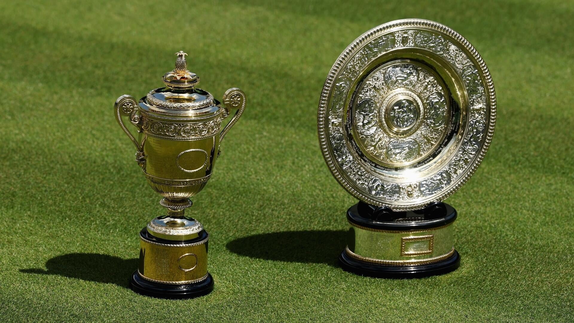 Two ornate silver tennis trophies, one a lidded cup and the other a decorative plate on a black base, rest on a grass court.