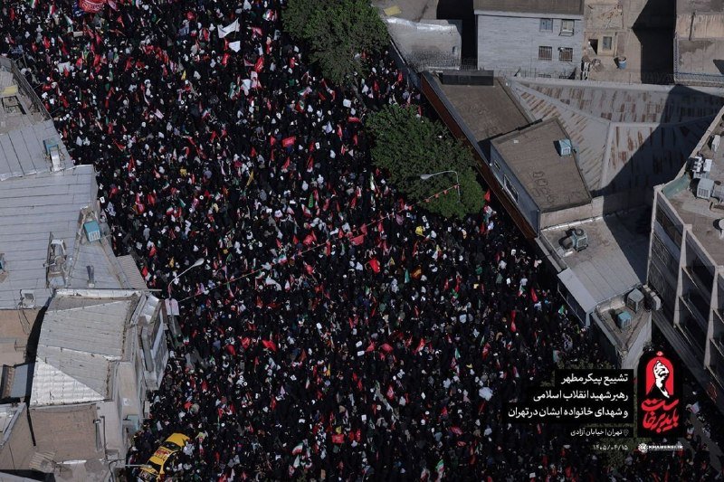 An aerial view shows a massive densely packed crowd waving numerous red, white, and black flags while filling a wide urban street flanked by multi-story buildings, with Persian text overlaid in the corner.