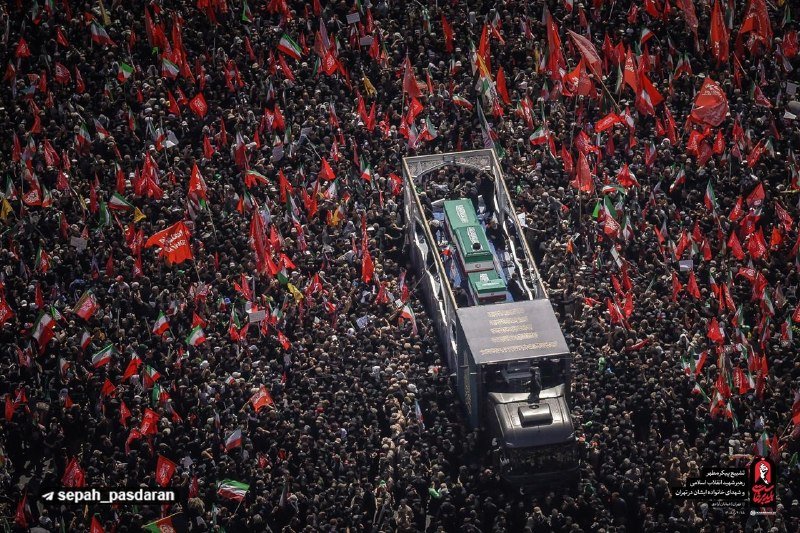 An aerial view shows a massive crowd waving red flags surrounding a flatbed truck carrying a casket, with a "sepah_pasdaran" watermark visible.