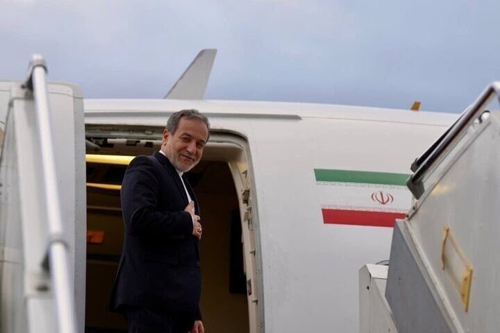 A bearded man in a dark suit smiles while standing at the open door of a white aircraft displaying the Iranian flag.