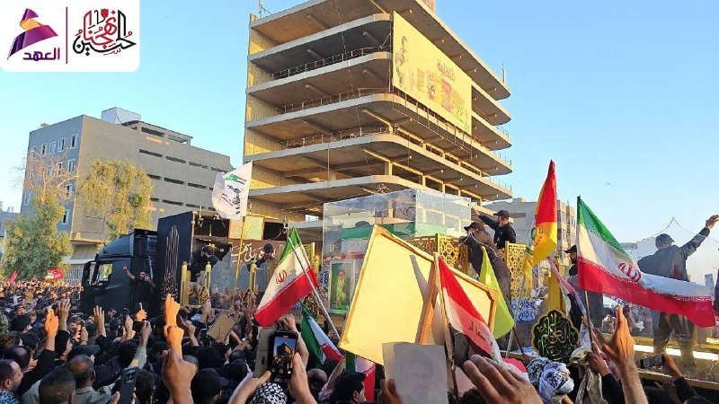A large crowd gathers outdoors waving Iranian flags and yellow banners in front of a coffin and an unfinished multi-story building under construction.