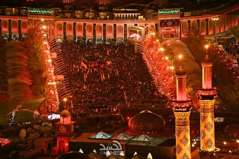 A large crowd gathers between golden-domed shrines and illuminated minarets at night, with arched walkways lit in red surrounding the gathering.