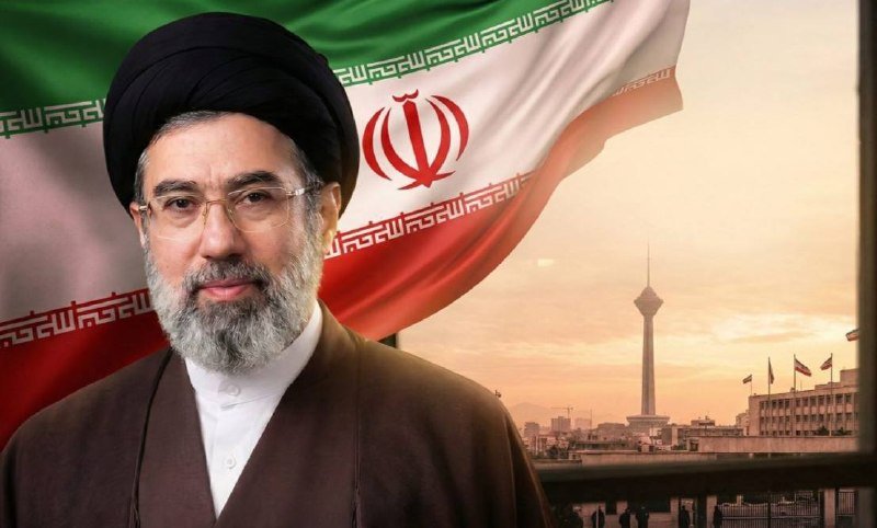 A man wearing a black turban and glasses is shown in front of the Iranian flag, with a city skyline and a tall tower visible in the background.