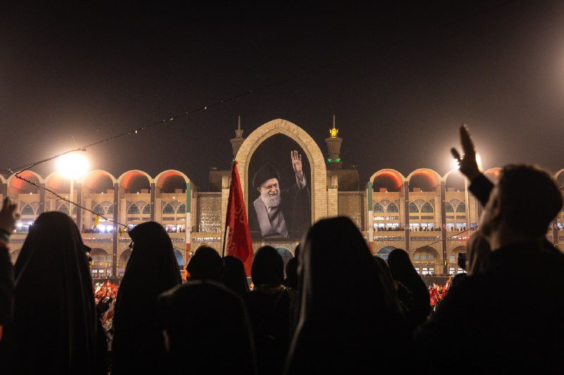 Silhouetted crowd raises hands at night before an illuminated archway displaying a waving portrait of a bearded cleric.
