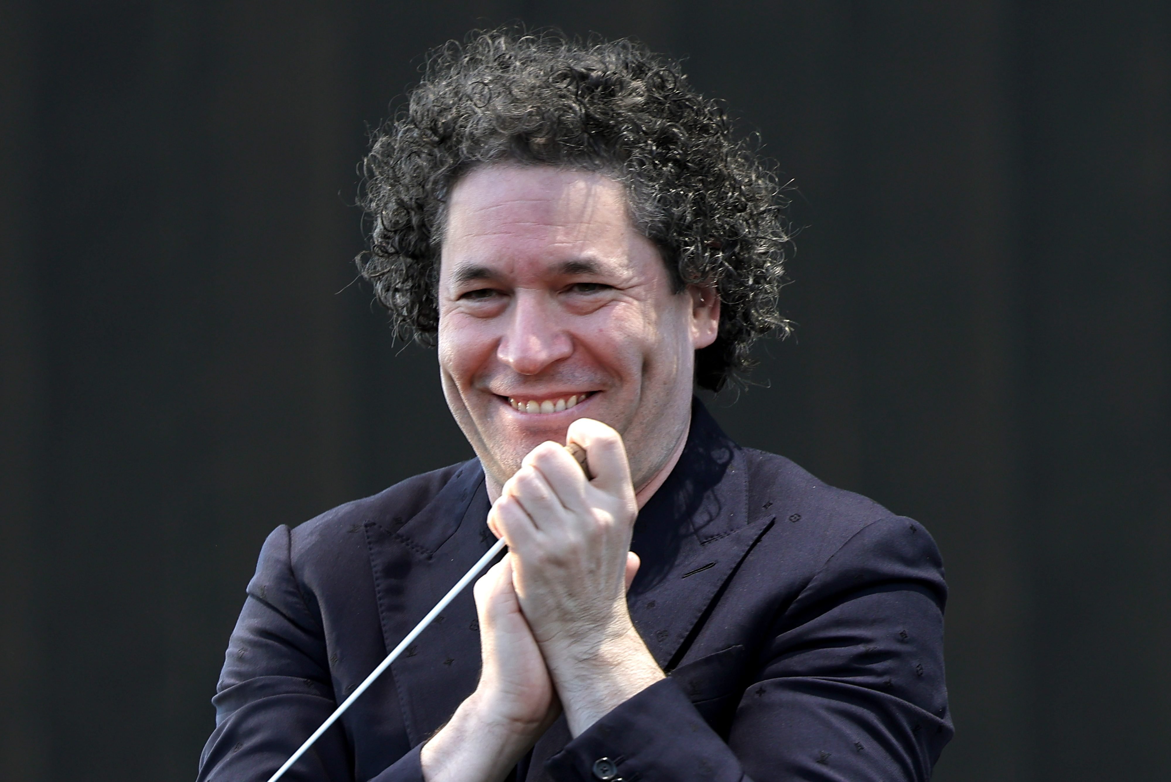 A curly-haired man in a dark navy jacket smiles while holding a conductor's baton near his chin against a black background.