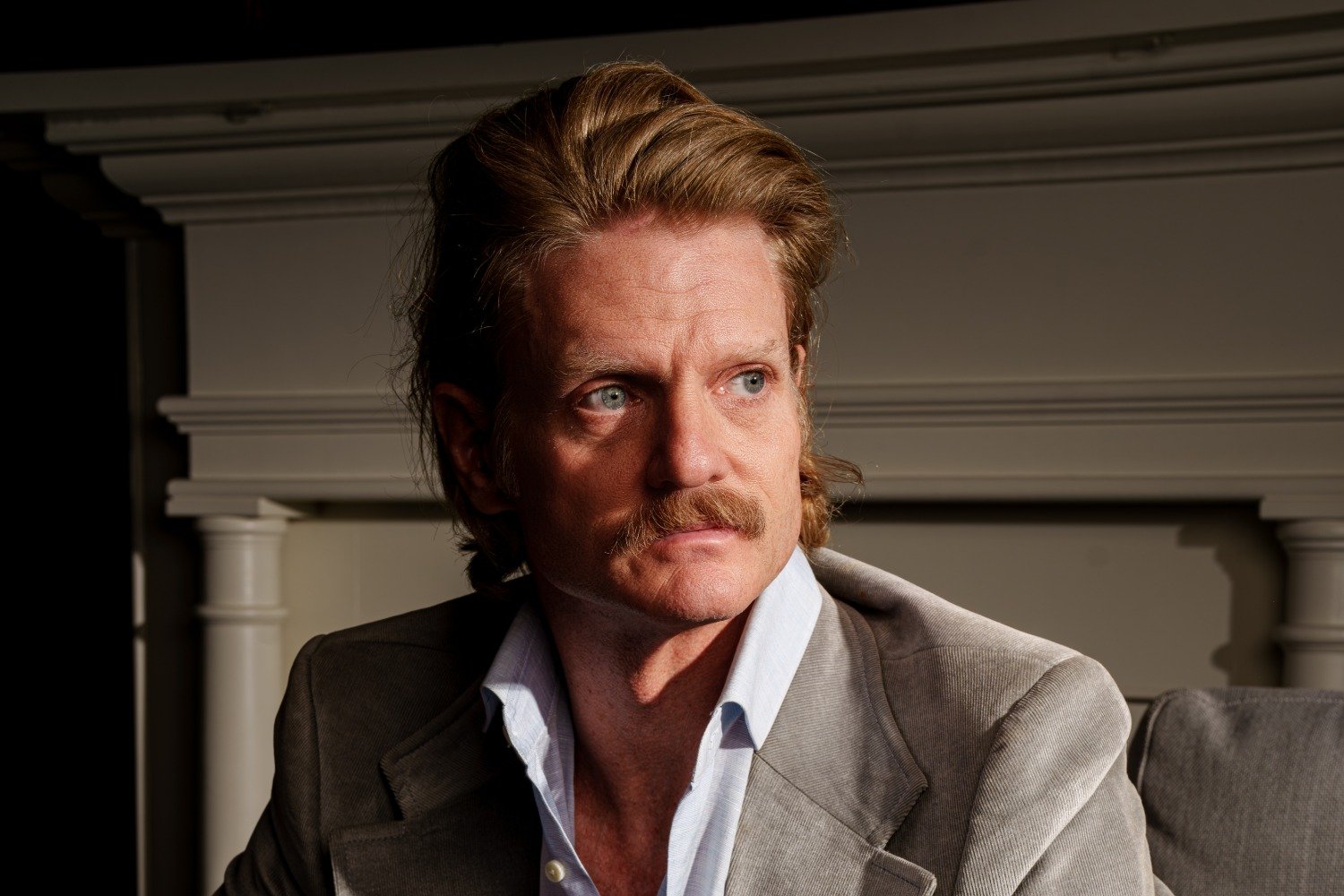 A man with a mustache and swept-back hair, wearing a beige blazer over an open-collared shirt, sits in dramatic lighting against a paneled wall.