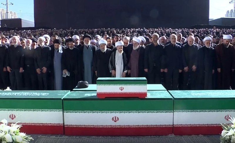 Clerics in white and black robes stand behind coffins draped in Iranian flags during an outdoor ceremony with a large crowd.
