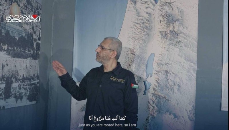 A bearded man in a dark shirt gestures toward a large map of the region displayed on the wall behind him, with Arabic and English subtitles visible below.