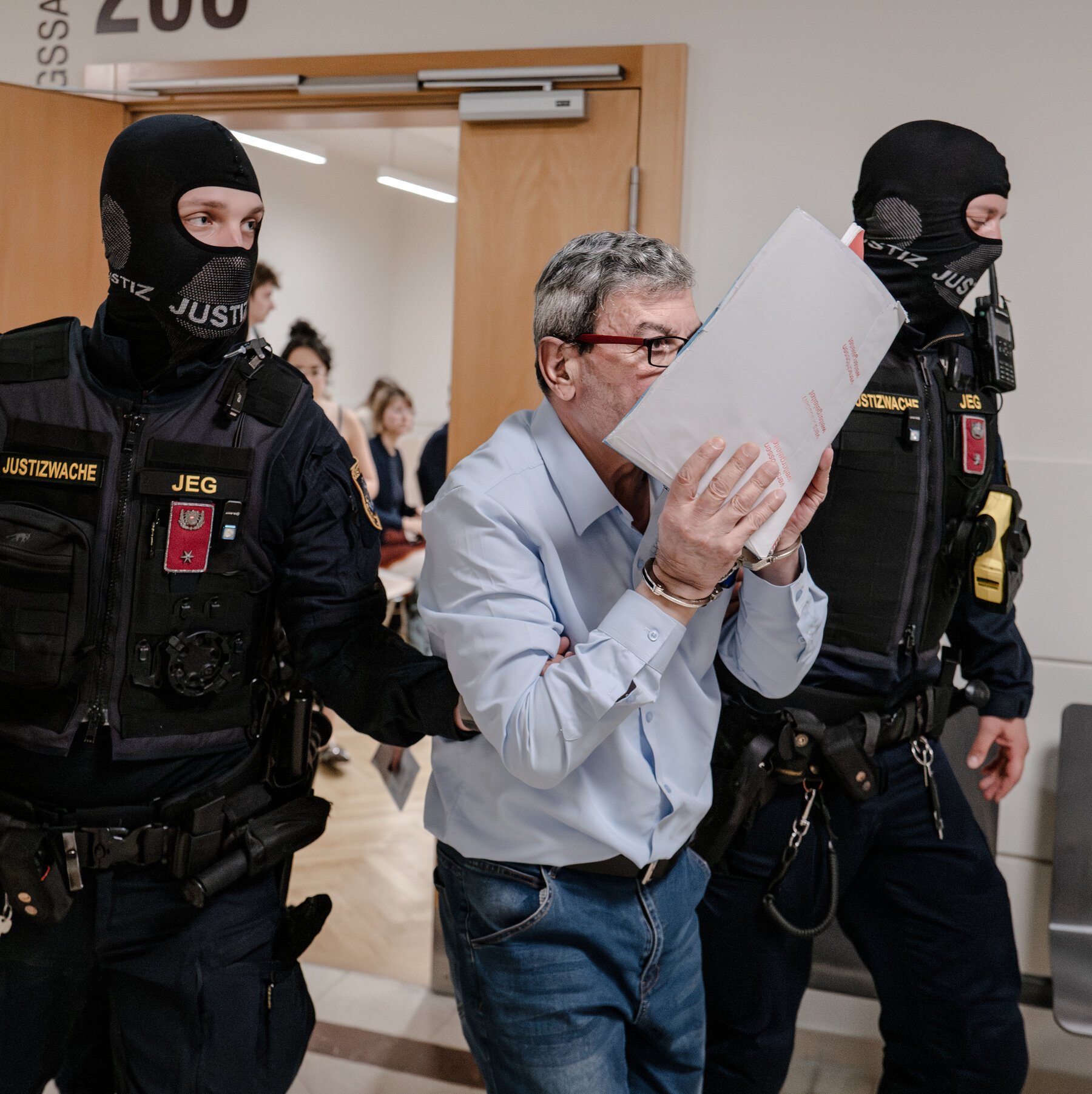 Two masked officers in dark uniforms labeled "JUSTIZWACHE JEG" escort a gray-haired man in handcuffs holding folders in front of his face through a doorway.