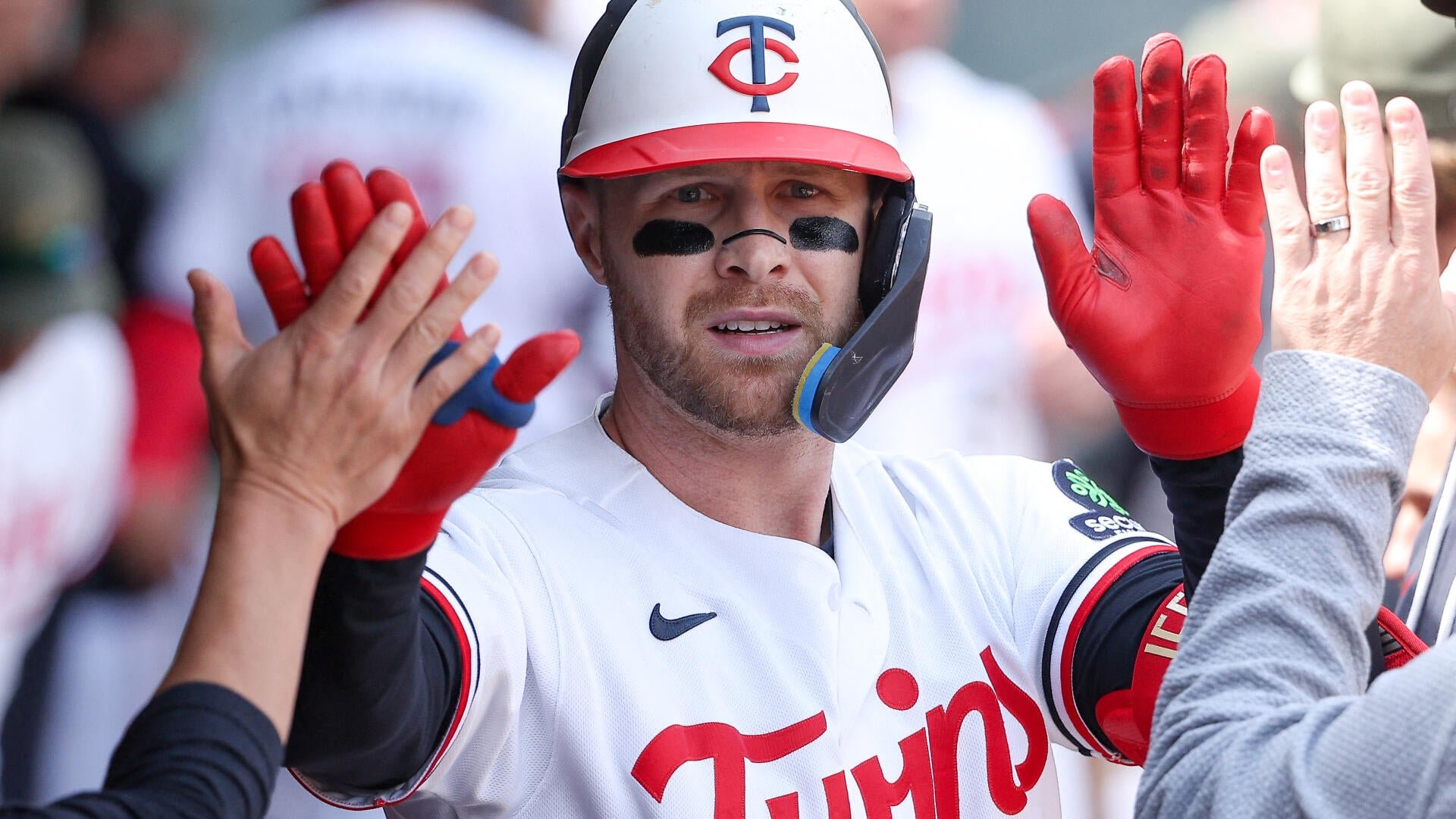 A baseball player in a white Twins uniform, batting helmet, and red gloves raises his hands for high-fives in a dugout.