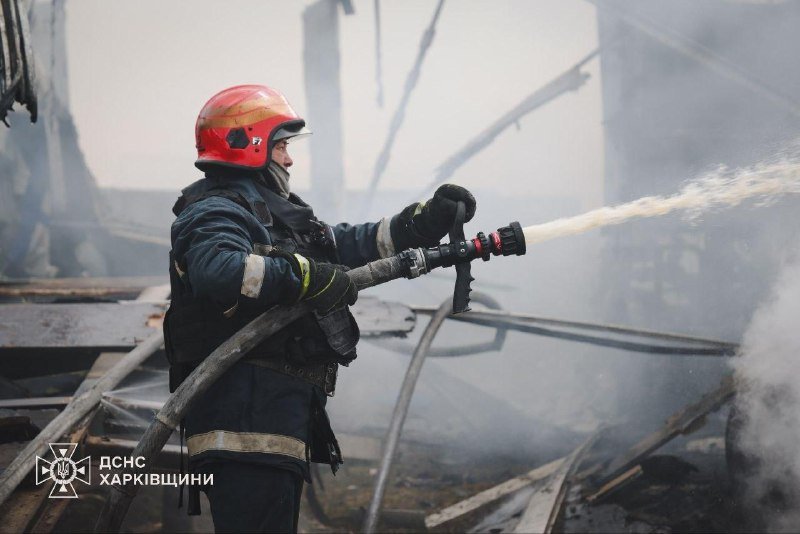 A firefighter in a red helmet sprays water from a hose amid smoldering, debris-filled ruins.