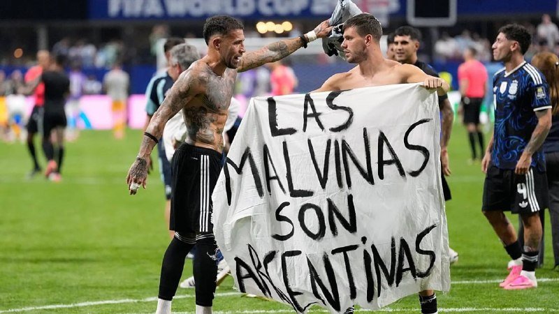 Two shirtless soccer players on a field hold up a banner reading "LAS MALVINAS SON ARGENTINAS" while a FIFA World Cup sign is visible in the background.