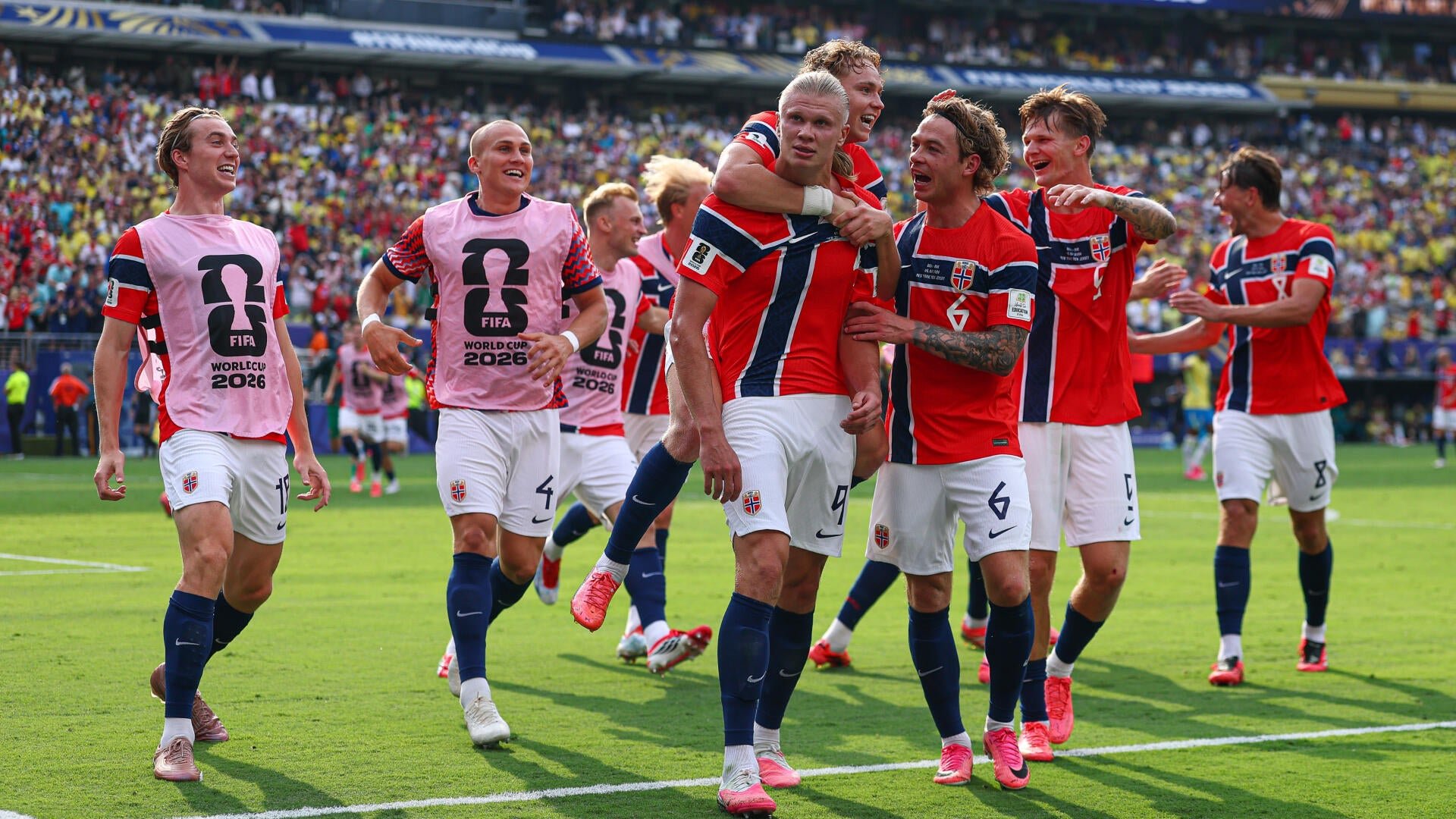 Football players in red, white, and blue uniforms celebrate on a stadium field, with teammates in pink bibs marked "FIFA World Cup 2026" running toward them as a player jumps onto another's back.