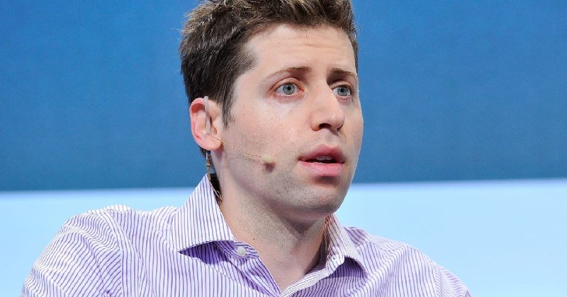 A man with short brown hair wearing a purple and white striped shirt speaks while wearing a headset microphone against a blue background.